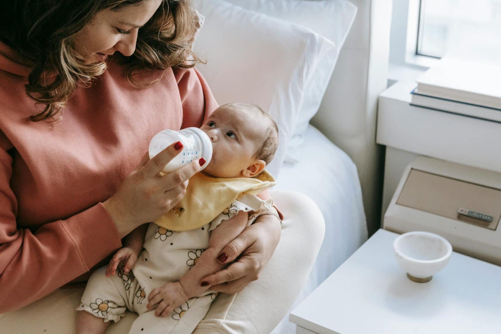 Signs Your Baby is Getting Enough Milk: A woman in a pink sweatshirt feeding milk from a bottle to a baby wearing a yellow bib and patterned clothing, while sitting on a bed next to a bedside table.