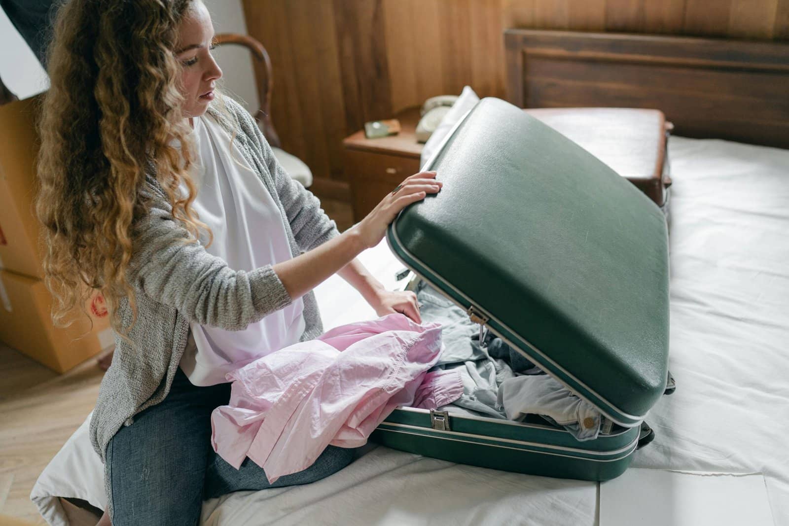 A woman with long curly hair packing clothes into a green suitcase on a bed.