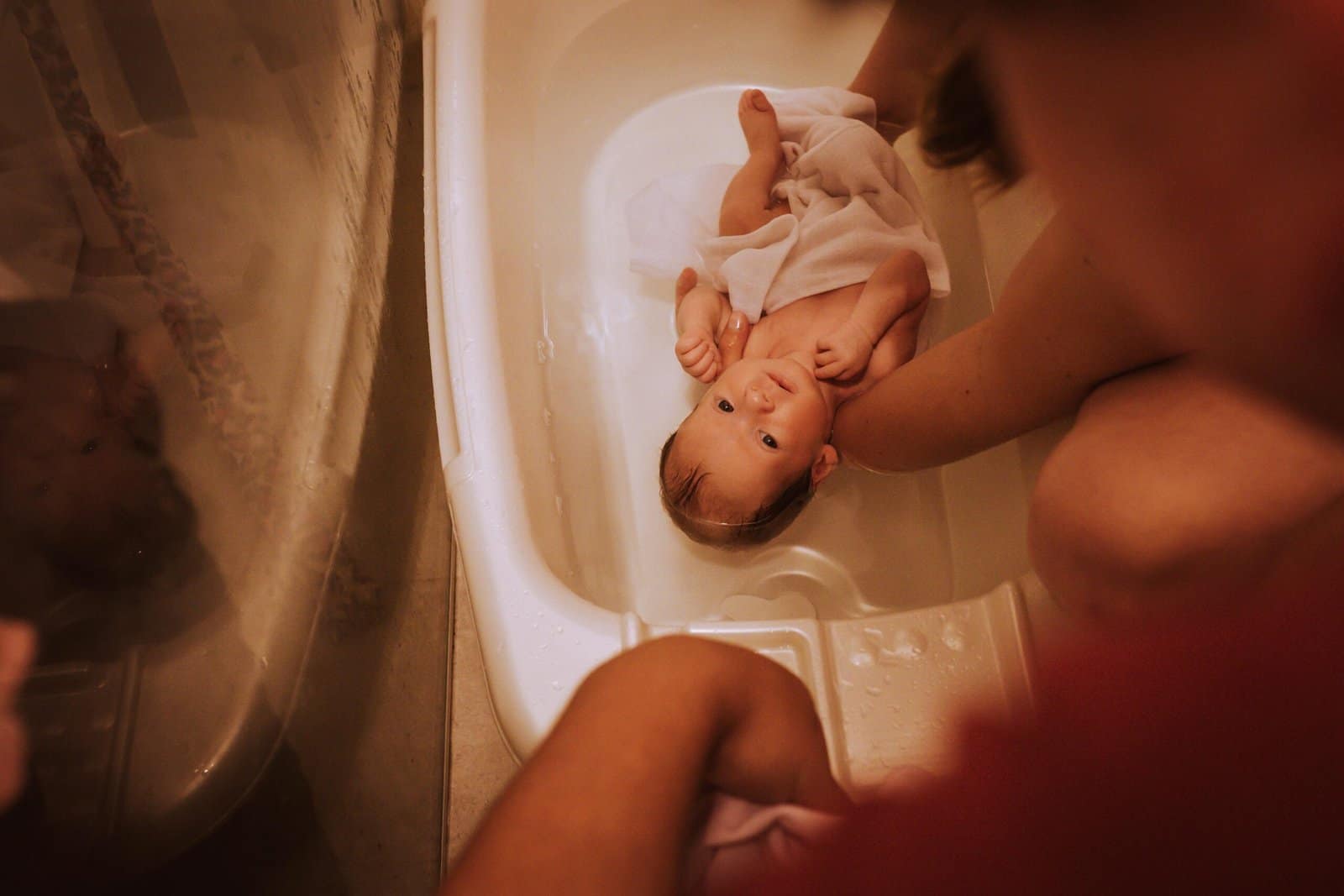 Newborn Bathing Guide: A baby lying in a small white bathtub with a towel partially covering them, surrounded by the arms of an adult providing support.