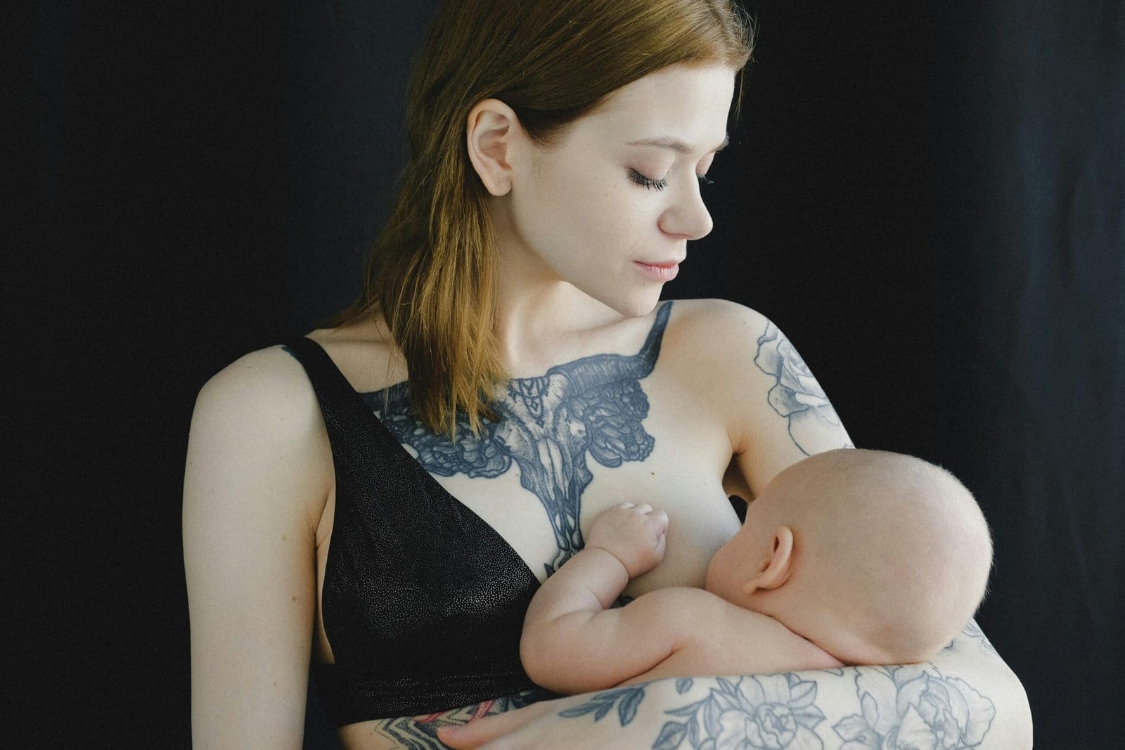 Breastfeeding Tips for New Moms: A woman with tattoos on her arms and chest is breastfeeding a baby against a dark background.