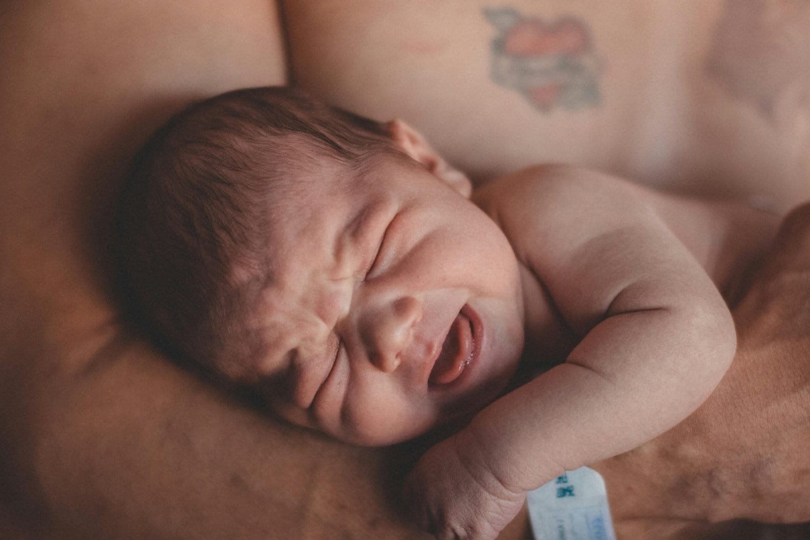 How to Soothe a Crying Baby: A newborn baby crying while being cradled in an adult's arms, with a tattoo visible on the adult's chest in the background.