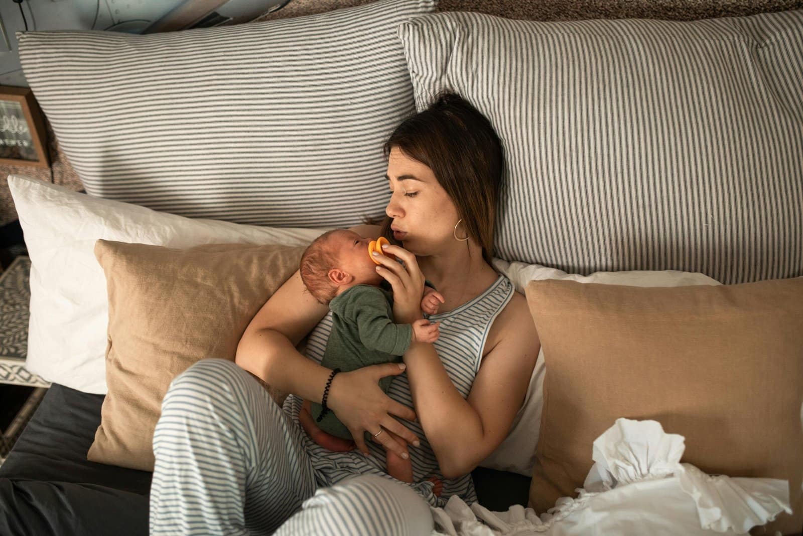 Postpartum Recovery in 2025 1 A woman in striped pyjamas is lying in bed, gently holding and feeding an infant with a bottle. They are surrounded by striped and plain pillows.