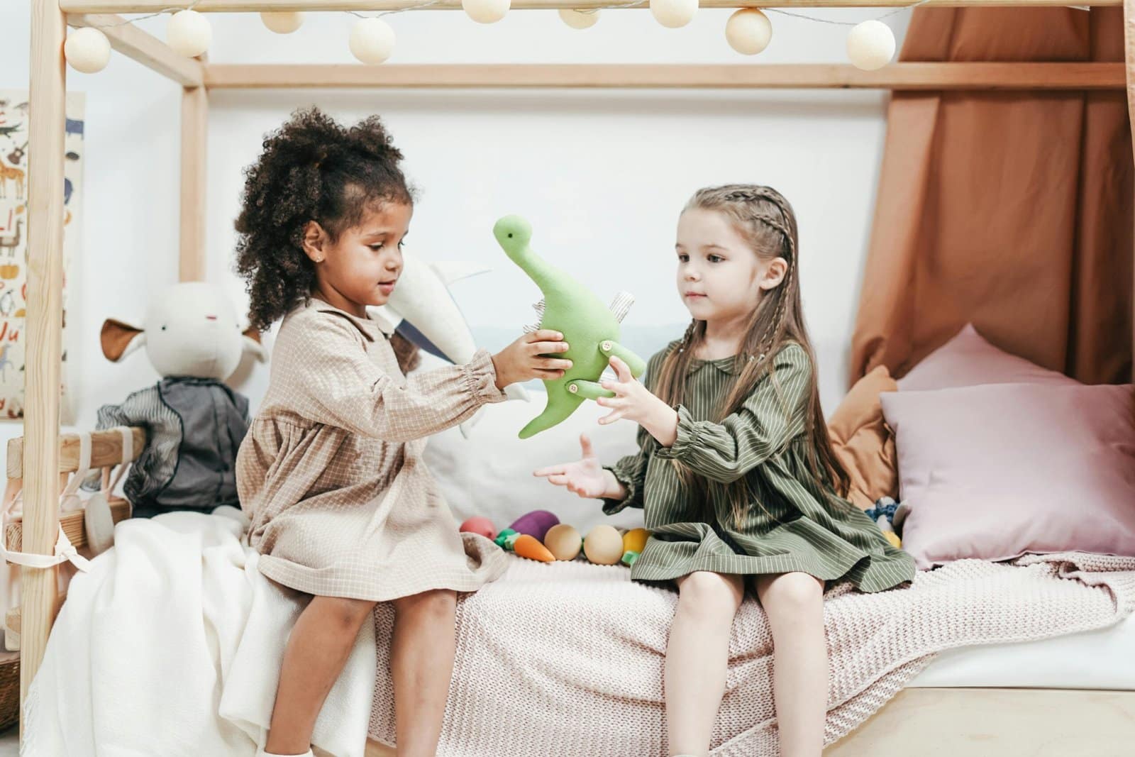 Sibling Room Sharing v Separate Bedrooms 1 Two young girls sitting on a bed, playing with a green dinosaur plush toy in a cozy room with soft lighting and pillows.