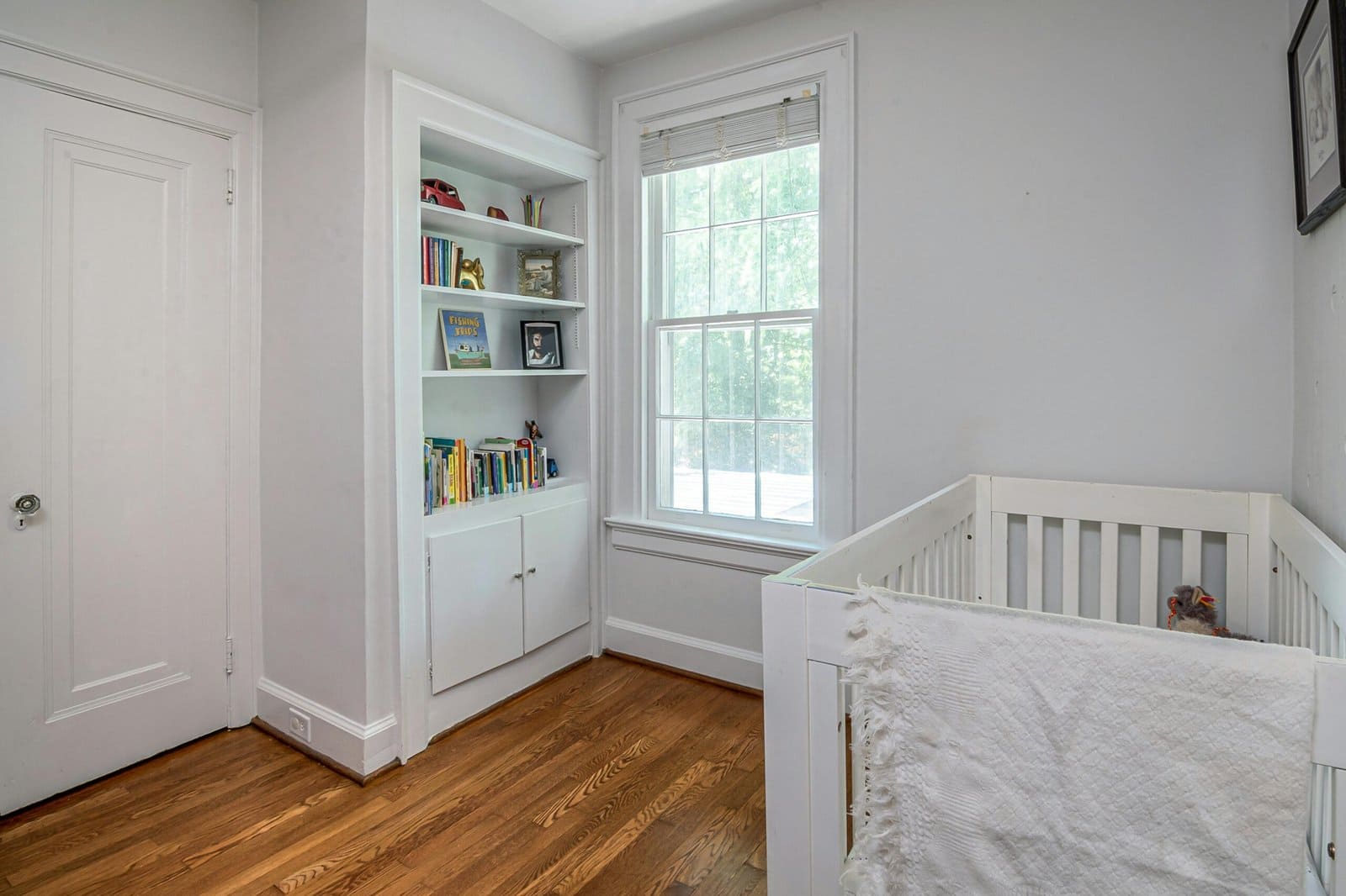 Your Guide to Safe Sleep for Baby (2025 Update) 2 A nursery with a white crib, a fluffy white blanket draped over the side, a built-in bookshelf filled with books and toys, a large window letting in natural light, and a closed white door.